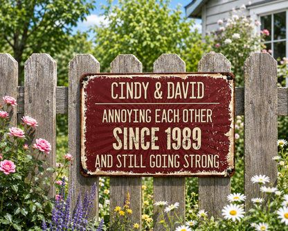 A rectangular, distressed burgundy metal sign with white text. The sign reads "Cindy & David Annoying Each Other Since 1989 And Still Going Strong." The sign is mounted on a weathered wooden fence, surrounded by flowers.