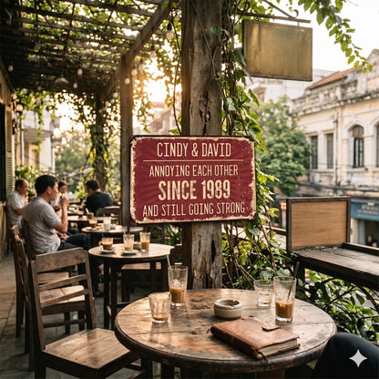 A burgundy metal sign with white text that reads "CINDY & DAVID ANNOYING EACH OTHER SINCE 1989 AND STILL GOING STRONG." The sign is mounted on a wooden post in an outdoor cafe setting.
