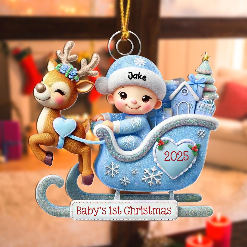 May include: A hanging Christmas ornament featuring a reindeer pulling a light blue sleigh with a baby wearing a matching hat labeled Jake. The sleigh holds gifts and a tree, with a heart displaying 2025 and a base reading Baby's 1st Christmas.