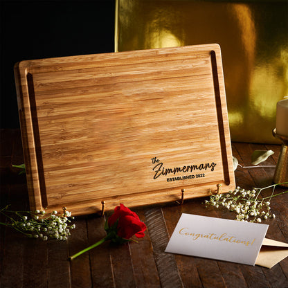 A rectangular bamboo cutting board with a juice groove along the perimeter. The board is engraved with the text "the Zimmermans ESTABLISHED 2022" in black lettering. It is displayed on a small stand with a red rose and a card.