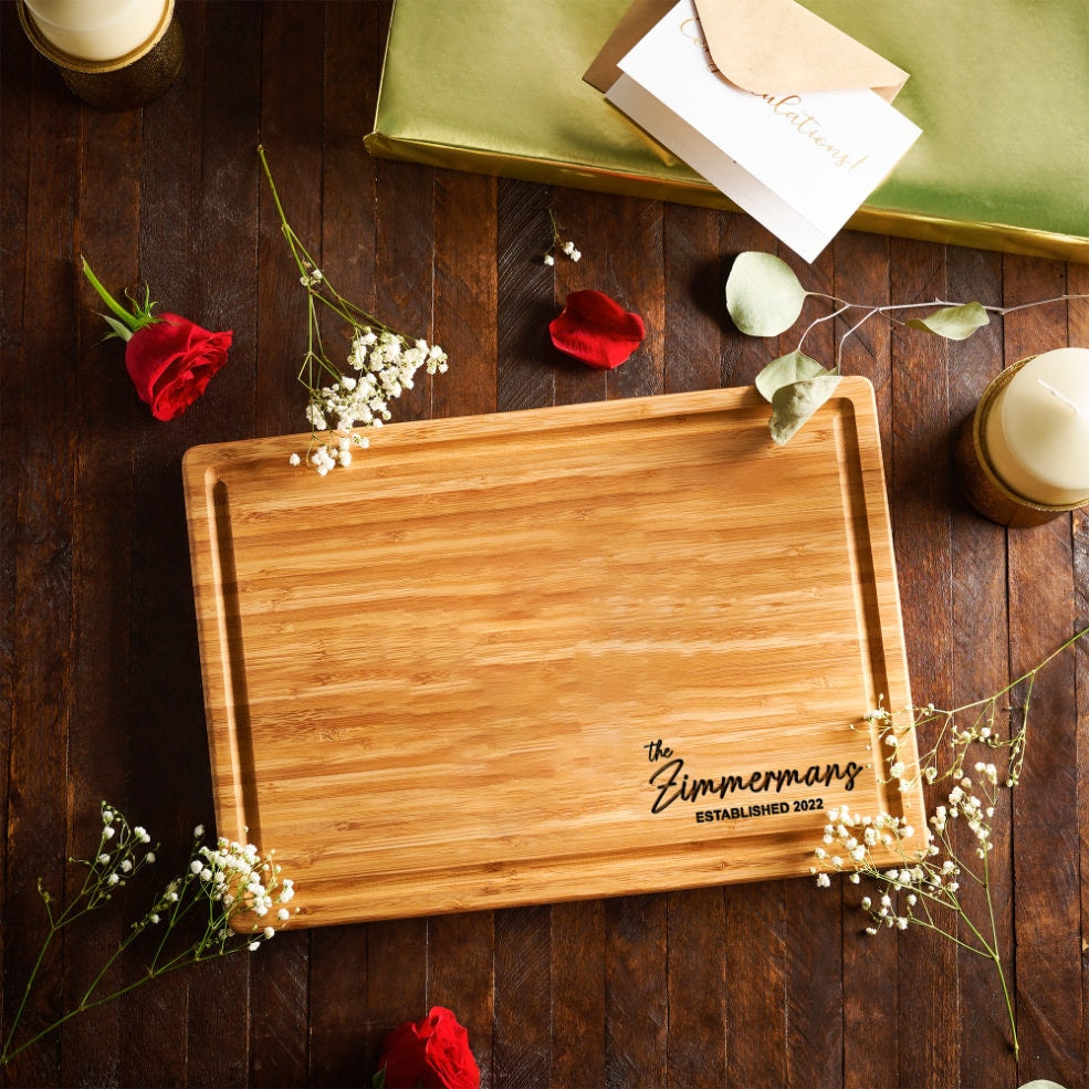 A rectangular bamboo cutting board with a juice groove along the perimeter. The board features black engraved text in the bottom right corner that reads, the Zimmermans ESTABLISHED 2022, in a combination of script and block fonts.