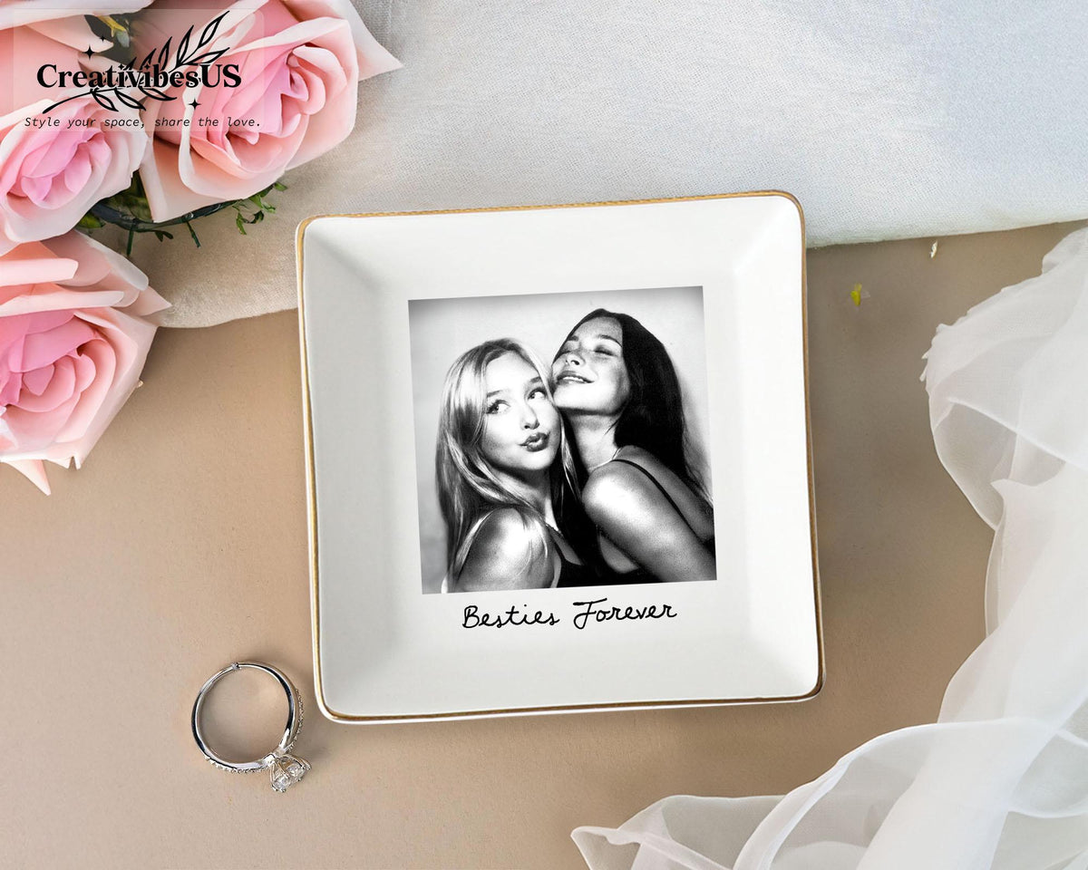 A square white ceramic trinket dish with a gold-toned rim. The center features a black and white photograph of two people with the text Besties Forever printed in black script below the image. A silver ring sits beside the dish.
