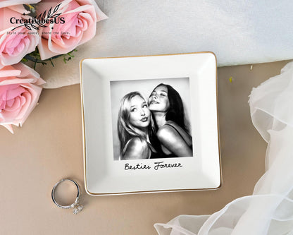 A square white ceramic trinket dish with a gold-toned rim. The center features a black and white photograph of two people with the text Besties Forever printed in black script below the image. A silver ring sits beside the dish.