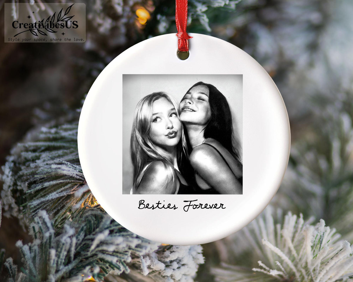 A round white ceramic ornament featuring a black and white square photograph of two people posing together. Below the photo, the text Besties Forever is printed in black script. A red ribbon is attached to the top for hanging.