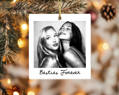 A square white ceramic ornament featuring a black and white photograph of two people posing together. The text Besties Forever is printed in black cursive script at the bottom of the ornament. It hangs from a gold string.