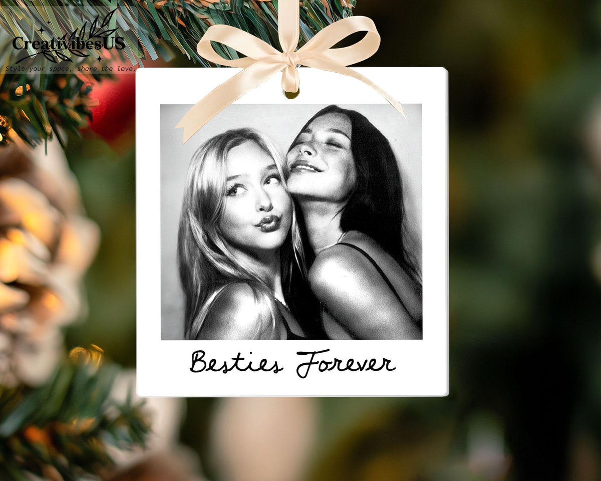 A square white hanging ornament featuring a black and white photograph of two individuals posing together. The ornament is topped with a light beige ribbon bow and includes the black cursive text Besties Forever at the bottom.