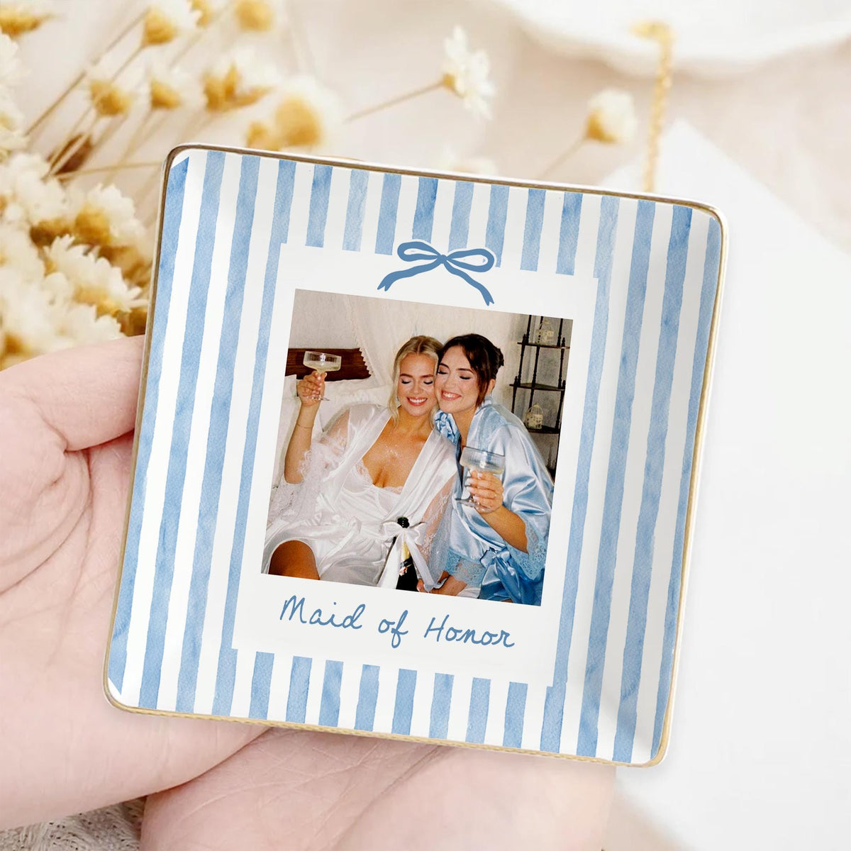 A square ceramic trinket dish featuring a light blue and white watercolor striped pattern. The center displays a photo of two individuals holding glasses, framed like a polaroid with a blue bow graphic and the text Maid of Honor.