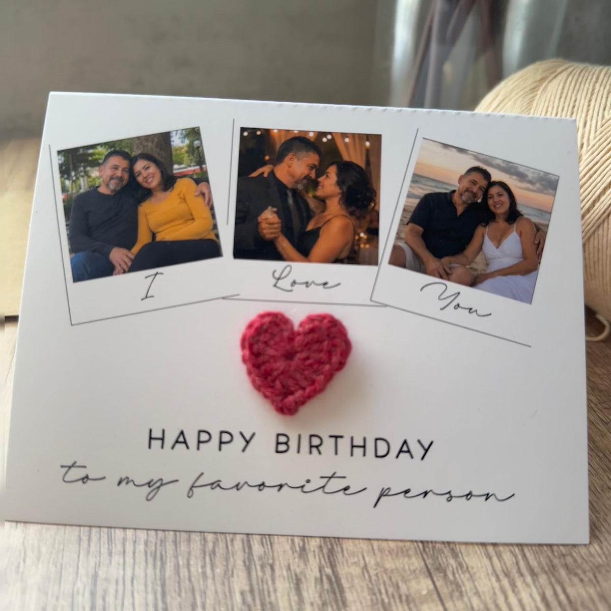 A white rectangular greeting card featuring three square photo frames with the text I Love You underneath them. A small red crocheted heart is attached to the center. The text HAPPY BIRTHDAY to my favorite person is printed below.