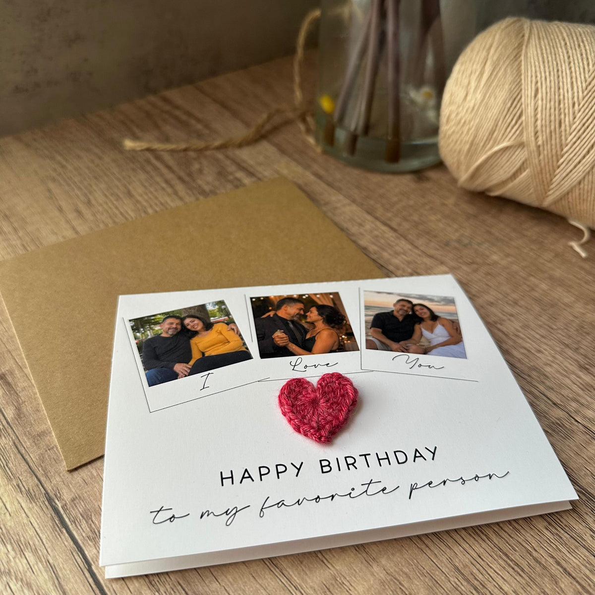 A white greeting card featuring three square photo prints of a couple, the handwritten text "I Love You", a small red crocheted heart, and the printed text "HAPPY BIRTHDAY to my favorite person" with a matching brown envelope.