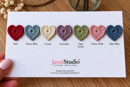 A white display card featuring seven small crocheted hearts in red, dusty blue, cream, lavender, sage green, dusty pink, and slate blue. The card includes the text JanaiStudio CUSTOM CREATIONS and a website URL at the bottom.