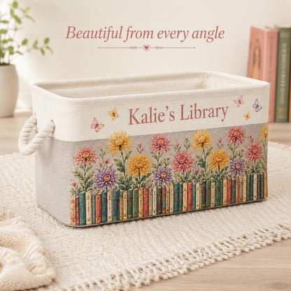 May include: A rectangular storage basket with cream and light gray fabric, featuring the text "Kalie's Library" in brown. The design includes a row of colorful books at the base, topped with pink, yellow, and purple flowers and butterflies.