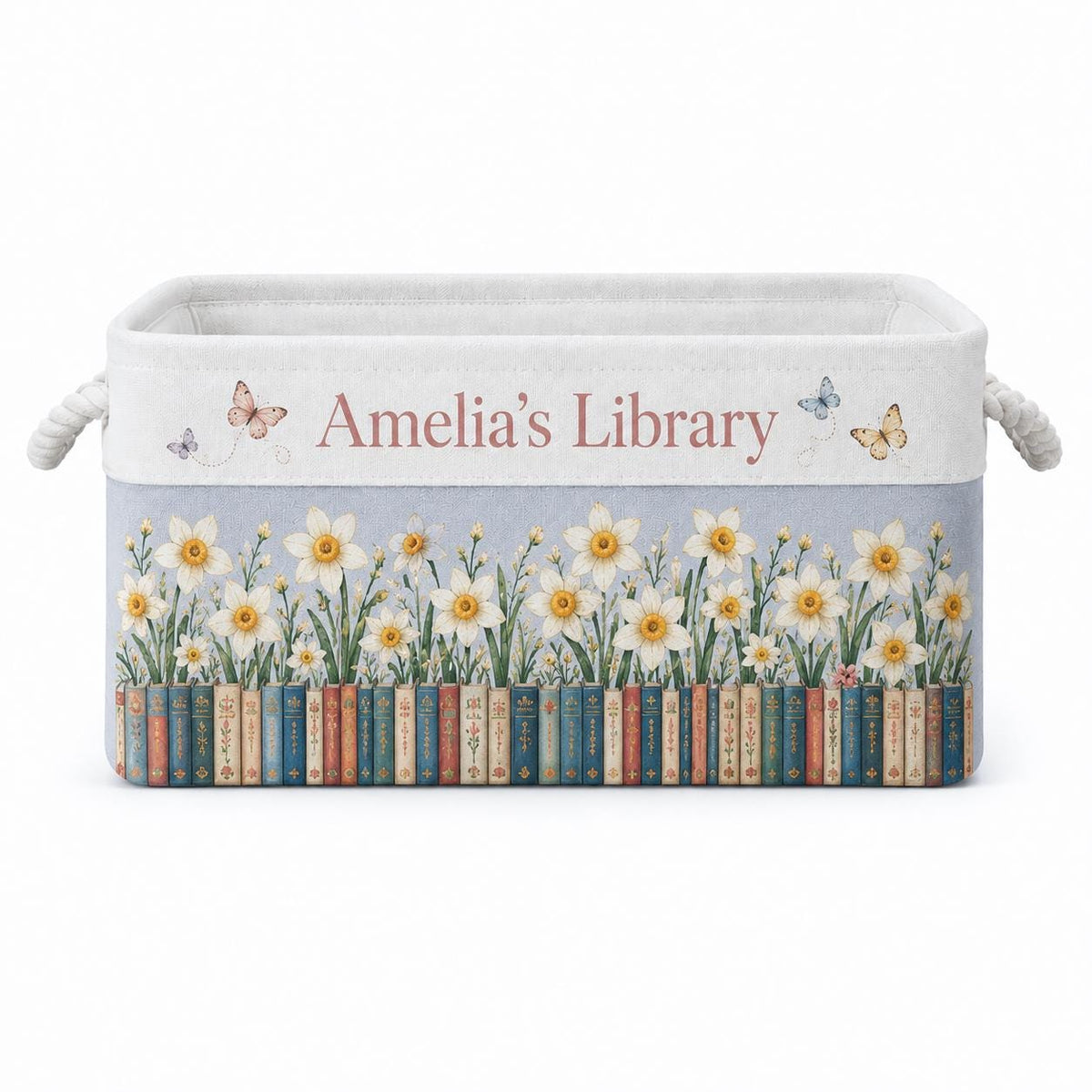 May include: A rectangular fabric storage bin with rope handles featuring the text Amelia's Library in pink. The design includes a row of colorful book spines at the base, topped with white daffodil flowers and small butterflies on a light blue background.