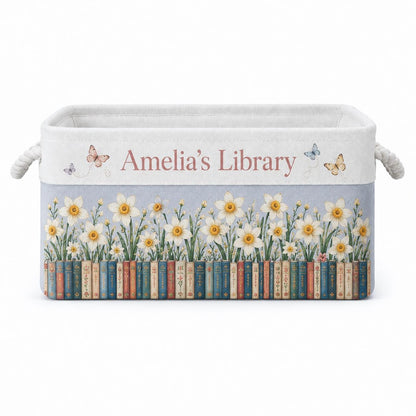 May include: A rectangular fabric storage bin with rope handles featuring the text Amelia's Library in pink. The design includes a row of colorful book spines at the base, topped with white daffodil flowers and small butterflies on a light blue background.