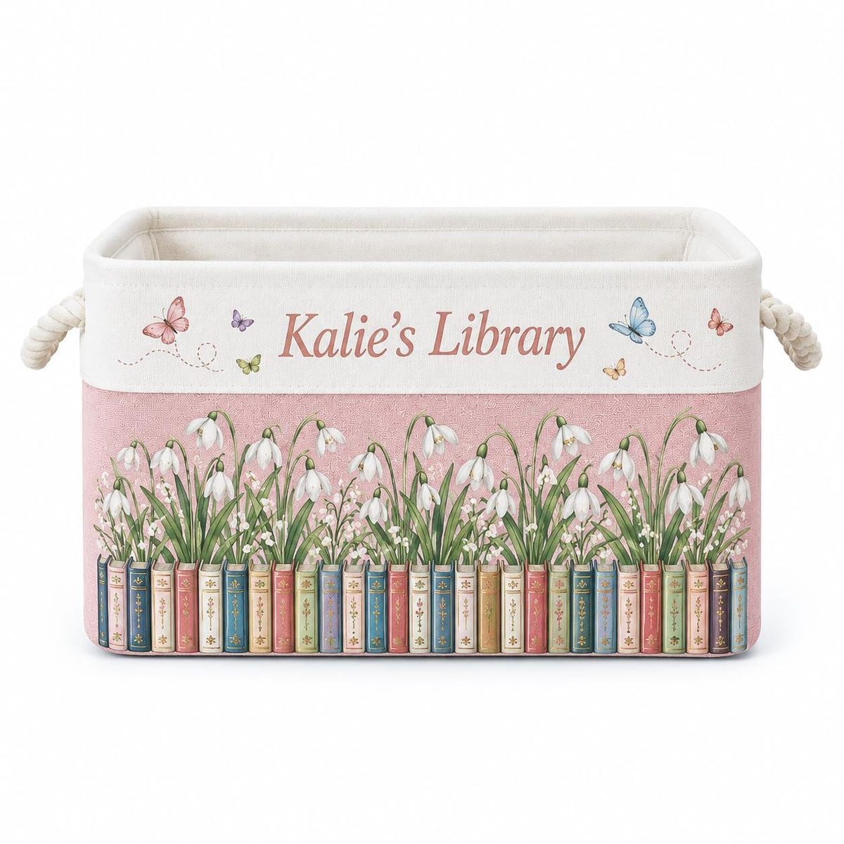 May include: A rectangular fabric storage bin with rope handles, featuring a cream top section with the text "Kalie's Library" and colorful butterflies. The pink lower section displays a row of books topped with white snowdrop flowers.