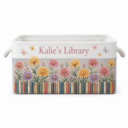 May include: A rectangular fabric storage bin with rope handles, featuring the text "Kalie's Library" in pink. The design includes a row of colorful books at the base, topped with a garden of yellow, pink, and purple chrysanthemums and butterflies.