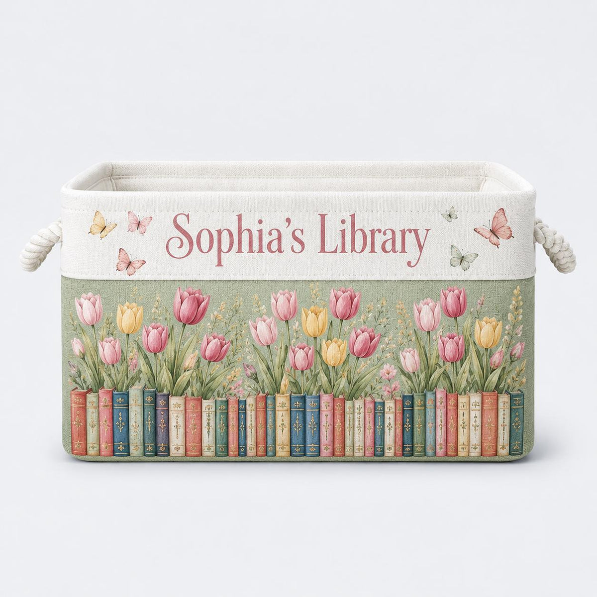 May include: A rectangular fabric storage bin with rope handles, featuring the text "Sophia's Library" in pink script. The design includes a row of colorful book spines at the base, topped with pink and yellow tulips and small butterflies.