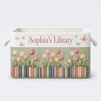 May include: A rectangular fabric storage bin with rope handles, featuring the text "Sophia's Library" in pink script. The design includes a row of colorful book spines at the base, topped with pink and yellow tulips and small butterflies.
