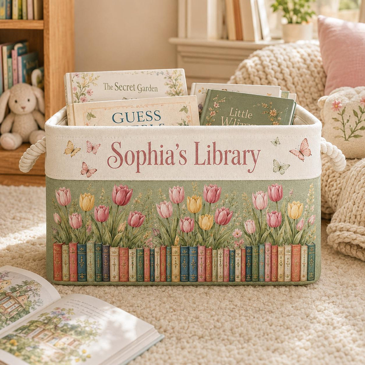 May include: A rectangular fabric storage bin with rope handles, featuring the text "Sophia's Library" in pink script. The design includes printed illustrations of colorful tulips and a row of books, accented with small butterflies on a cream background.
