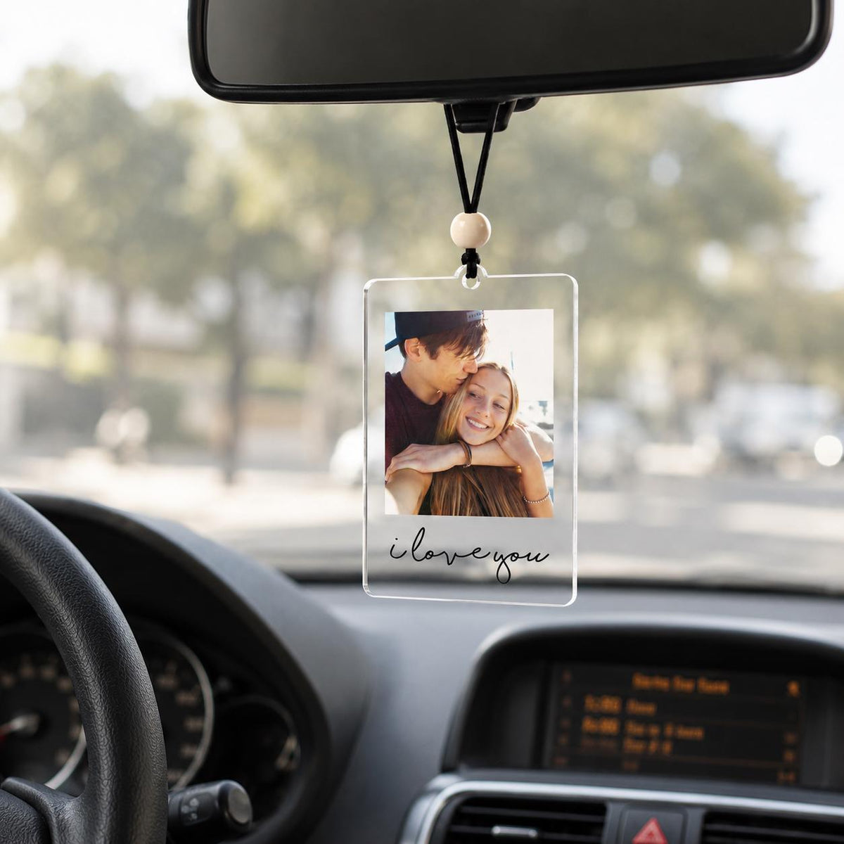 A rectangular clear acrylic car hanging ornament featuring a printed photograph of two people embracing. The ornament is suspended by a black cord with a single wooden bead and displays the black cursive text i love you.