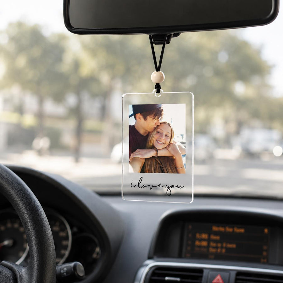 A rectangular clear acrylic car hanging ornament featuring a printed photograph of two people embracing. The ornament is suspended by a black cord with a single wooden bead and displays the black cursive text i love you.