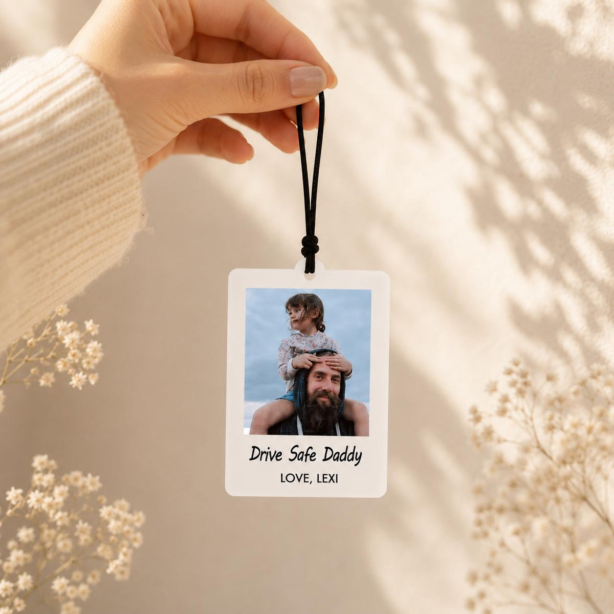 A rectangular white air freshener hanging from a black elastic cord. The center features a color photograph of a child sitting on a person's shoulders. Below the photo, the text reads Drive Safe Daddy and LOVE, LEXI in black.
