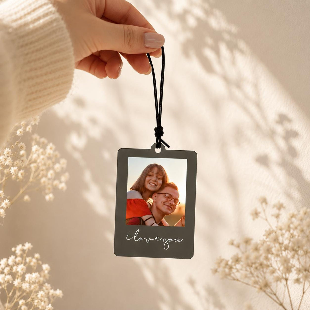 A rectangular hanging ornament with a dark gray border and a white printed script that reads "i love you." The center features a color photograph of two people embracing outdoors, suspended by a thin black elastic cord.