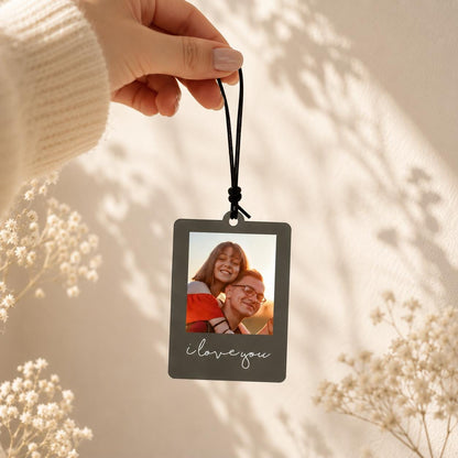 A rectangular hanging ornament with a dark gray border and a white printed script that reads "i love you." The center features a color photograph of two people embracing outdoors, suspended by a thin black elastic cord.