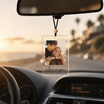 A rectangular clear acrylic car rearview mirror hanging ornament featuring a printed photograph of two people embracing. The ornament is suspended by a black cord with a single wooden bead and includes the text i love you in black.