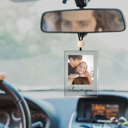 A rectangular clear acrylic car hanging ornament featuring a printed photograph of a couple embracing. The ornament is suspended by a black cord with a single light-colored wooden bead and displays the black cursive text i love you.