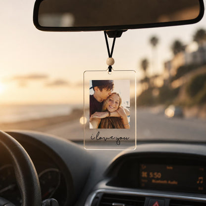 A rectangular clear acrylic car rearview mirror hanging ornament featuring a printed photograph of two people embracing. The ornament is suspended by a black cord with a single wooden bead and includes the text i love you in black.