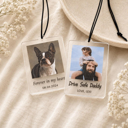 Two rectangular clear acrylic hanging ornaments with black cords. One features a Boston Terrier photo with the text "Forever in my heart" and "08.06.2024". The other shows a child on a man's shoulders with "Drive Safe Daddy" and "LOVE, LEXI".
