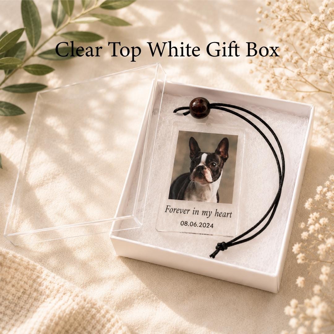 A white gift box with a clear lid containing a rectangular acrylic ornament. The ornament features a photograph of a black and white dog, the text "Forever in my heart," and the date "08.06.2024," attached to a black cord.