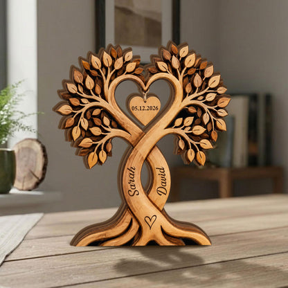 May include: A wooden tree sculpture with intertwined trunks, forming a heart shape. The sculpture features intricate leaf details and the names "Sarah" and "David" carved into the trunks. A heart-shaped tag displays the date "05.12.2026".
