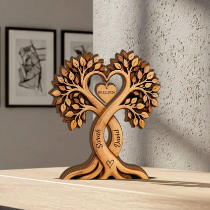 May include: A wooden tree sculpture with intertwined trunks, forming a heart shape at the top. The sculpture features engraved names "Sarah" and "David", a heart symbol, and the date "05.12.2026". The tree has detailed leaf carvings and a natural wood finish.