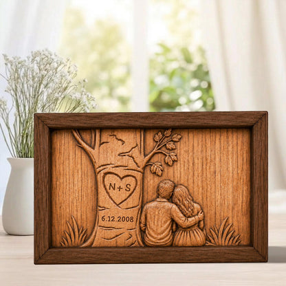 May include: A framed wooden carving depicting a couple embracing under a tree. The tree has a heart carved into it with the initials "N + S" and the date "6.12.2008". The frame is dark brown, and the carving is a lighter wood color.