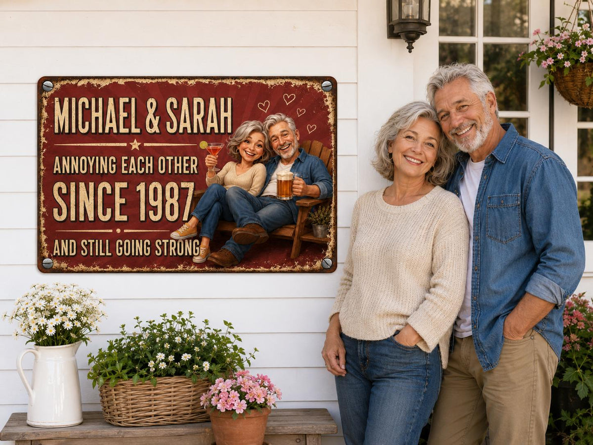 A rectangular metal sign with a distressed red background featuring the text MICHAEL & SARAH ANNOYING EACH OTHER SINCE 1987 AND STILL GOING STRONG. It depicts a cartoon couple sitting on a wooden chair with drinks and heart icons.