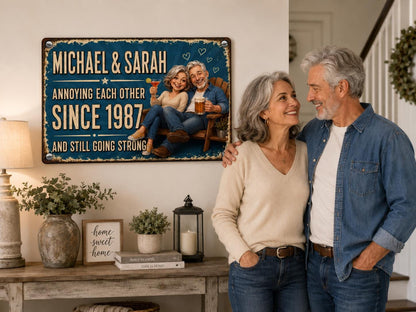 A rectangular metal sign with a blue background featuring the text MICHAEL & SARAH ANNOYING EACH OTHER SINCE 1987 AND STILL GOING STRONG. The sign displays a cartoon illustration of a couple sitting in a wooden chair with drinks.