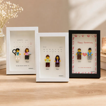 Three rectangular shadow box frames containing custom minifigures. Two white frames display calendar dates with names Saul, Mayra, Krish, and Riya. One black frame features the text Happy Anniversary with names Jason and Layla.