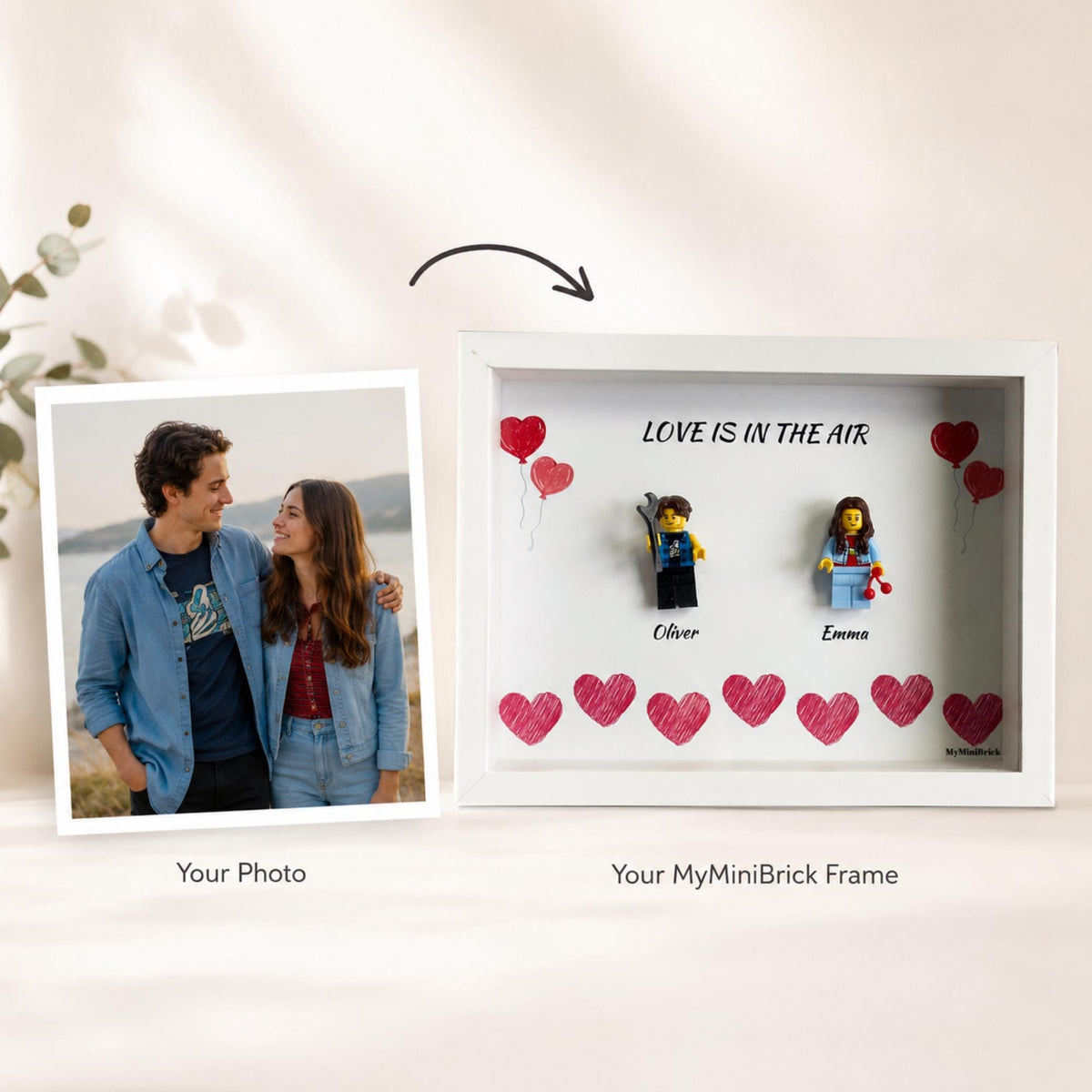 A white shadow box frame featuring two plastic minifigures, Oliver and Emma, with the text LOVE IS IN THE AIR and hand-drawn red hearts. Beside the frame is a square photograph of a couple, labeled as Your Photo.