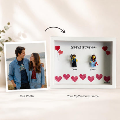 A white shadow box frame featuring two plastic minifigures, Oliver and Emma, with the text LOVE IS IN THE AIR and hand-drawn red hearts. Beside the frame is a square photograph of a couple, labeled as Your Photo.