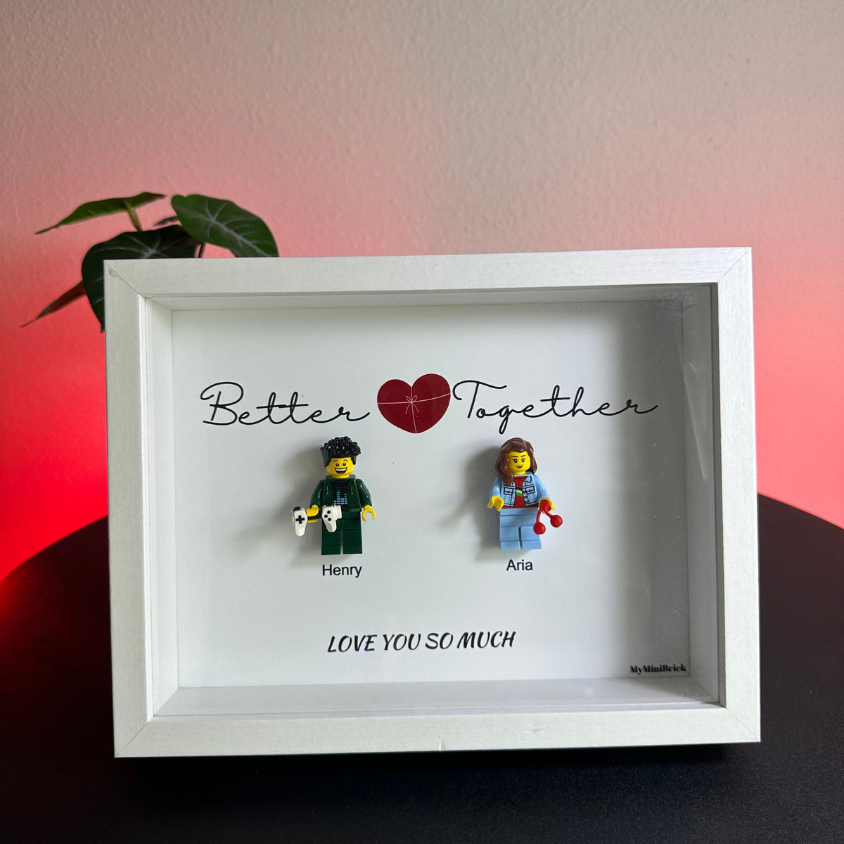 A white shadow box frame containing two minifigures. The text reads Better Together with a red heart, Henry, Aria, and LOVE YOU SO MUCH. One minifigure holds a white game controller, the other holds two red cherries. MyMiniBrick is printed.