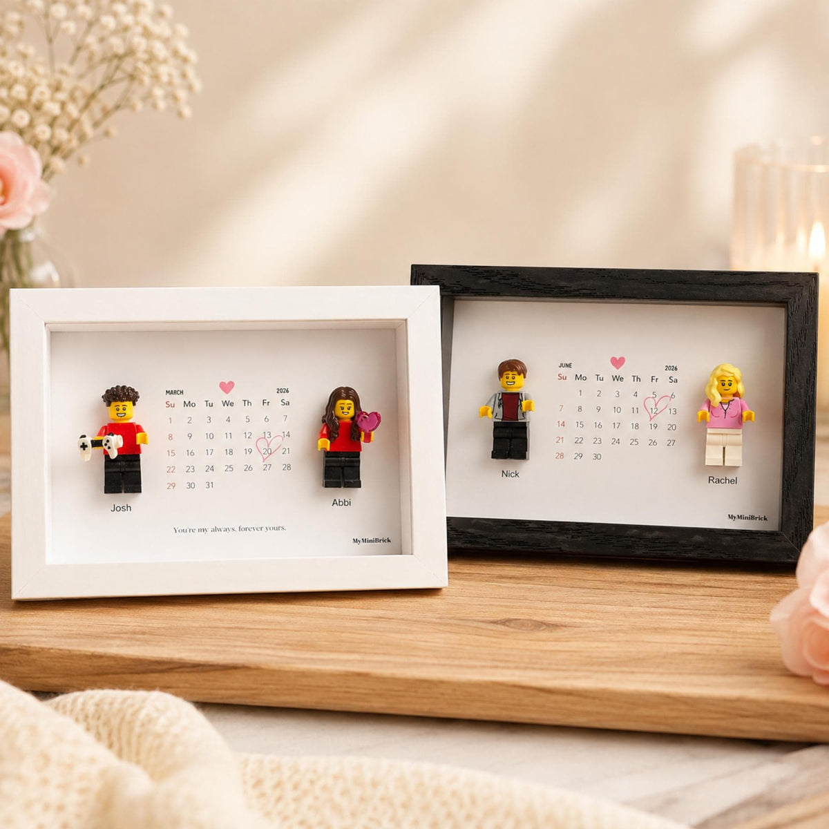 Two rectangular frames, one white and one black, each containing a personalized calendar for 2026 with two plastic toy figures. The white frame features Josh and Abbi with a March calendar, while the black frame shows Nick and Rachel with a June calendar. Each includes a small pink heart icon and a hand-drawn heart around a specific date.