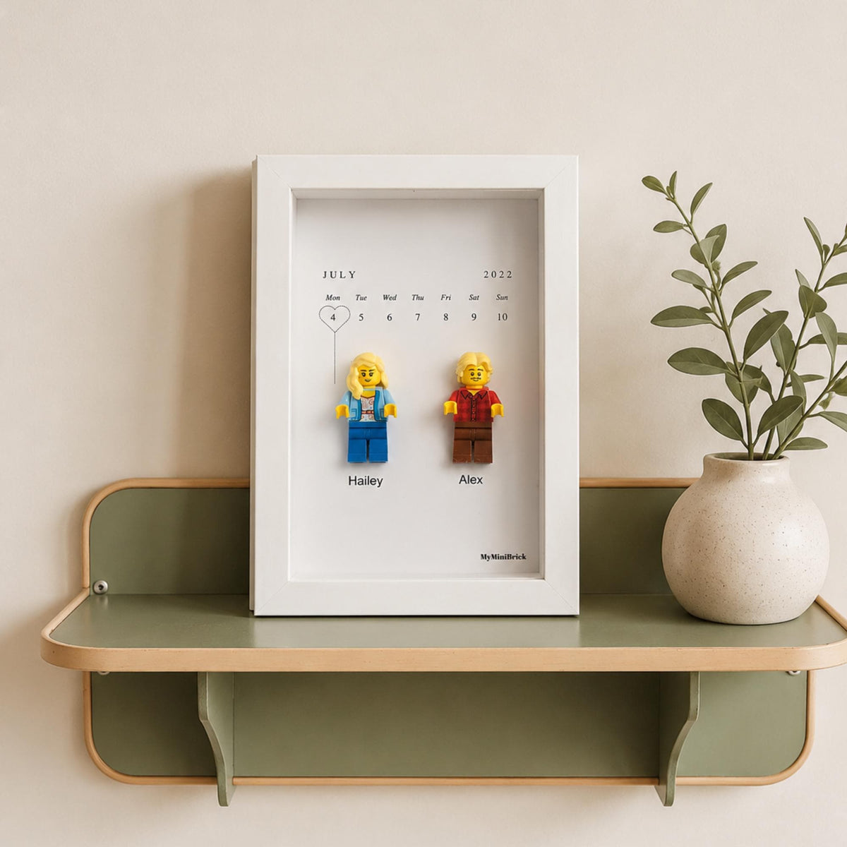 A white rectangular shadow box frame containing a July 2022 calendar print with two plastic toy figures attached. The figures are labeled Hailey and Alex. The text MyMiniBrick is printed at the bottom right of the white background.