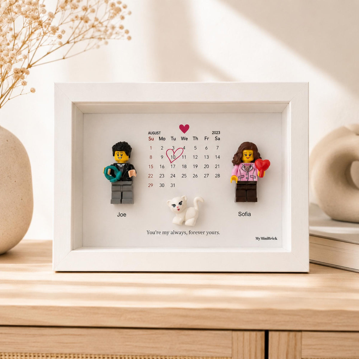 A white shadow box frame containing a calendar for August 2023 with a red heart around the 10th. Two plastic minifigures, Joe and Sofia, stand on either side of a small white cat figurine. The text reads, You're my always, forever yours.