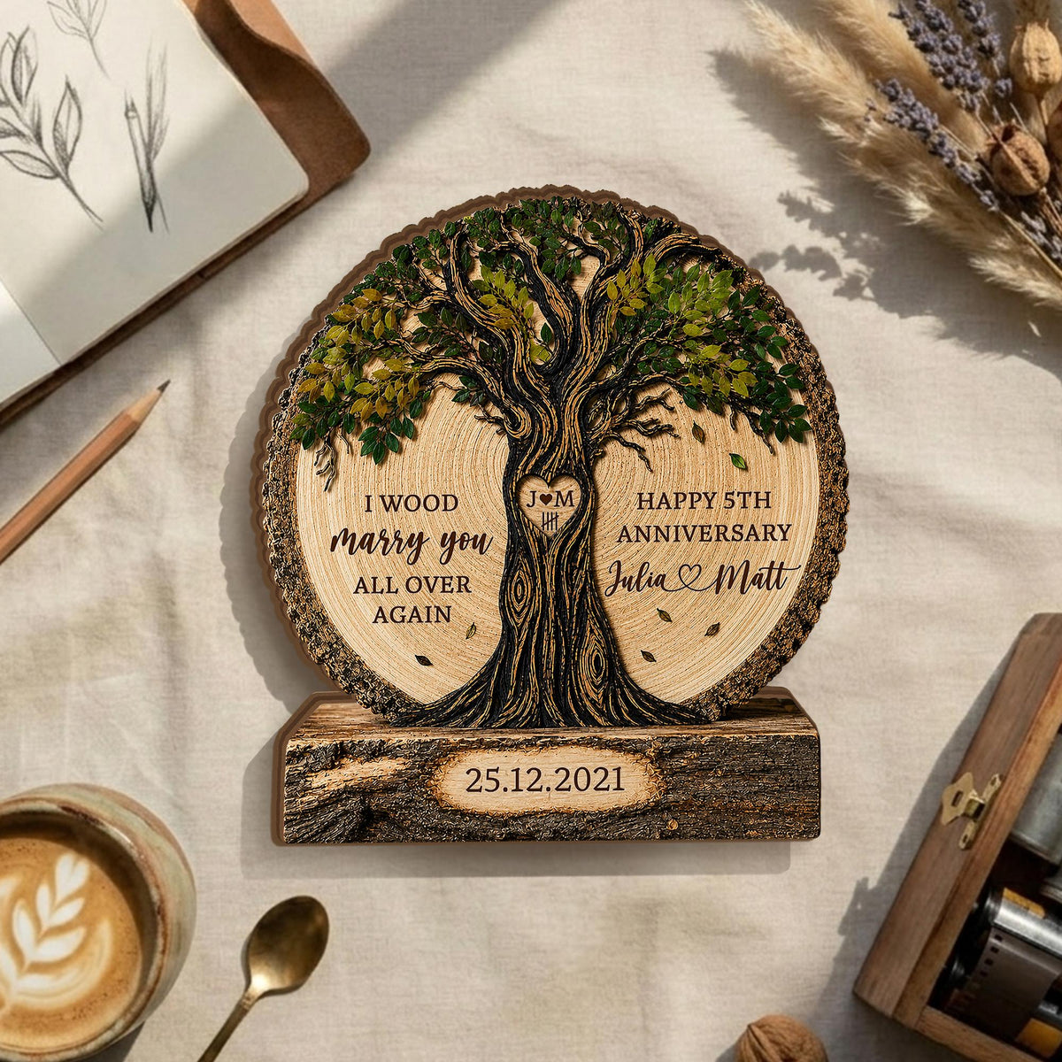May include: A circular wooden plaque featuring a laser-engraved tree with green foliage. The text reads I WOOD marry you ALL OVER AGAIN, HAPPY 5TH ANNIVERSARY, Julia and Matt, and the date 25.12.2021. A heart with J M is carved in the trunk.