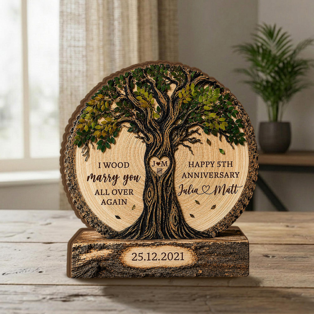 May include: A circular wooden plaque featuring a carved tree with green leaves. The text reads: I WOOD marry you ALL OVER AGAIN, HAPPY 5TH ANNIVERSARY, Julia and Matt, and 25.12.2021. A heart with J M is carved into the tree trunk on a base.