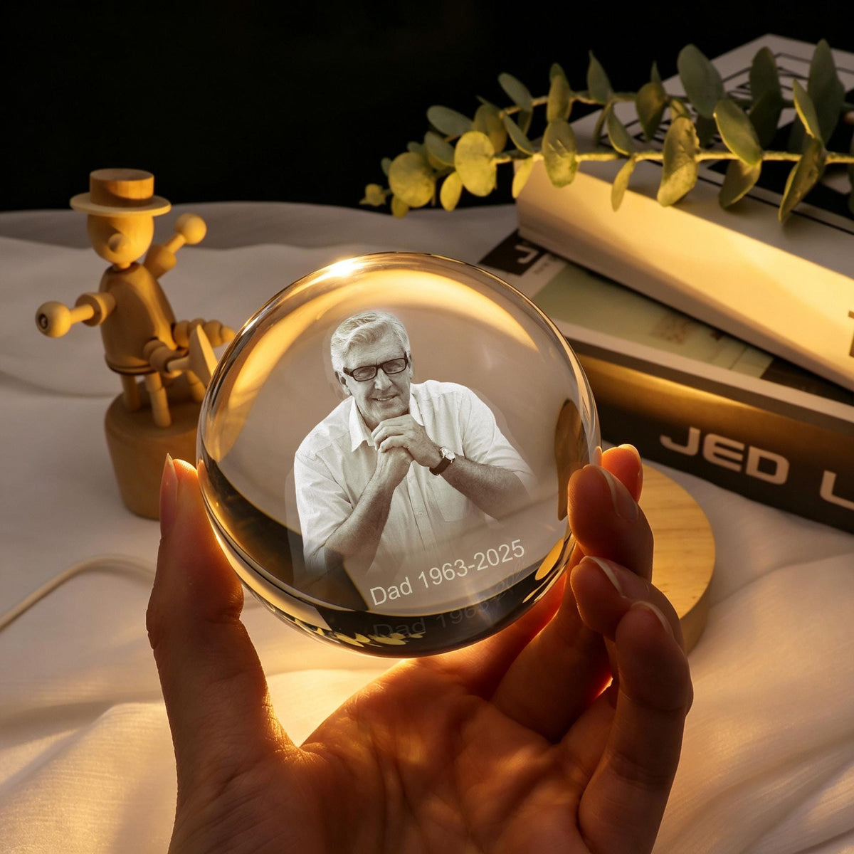 A clear glass sphere featuring a laser-etched 3D portrait of a person wearing glasses and a collared shirt, with the text "Dad 1963-2025" engraved at the bottom. The sphere is illuminated from below, highlighting the detailed image inside.