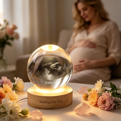 A clear glass sphere containing a 3D laser-etched ultrasound image of a fetus, resting on a circular wooden base. The base is illuminated from within and features the engraved text "Waiting For You" and "Baby X".