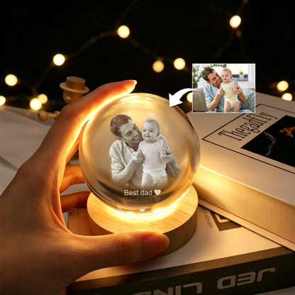 A clear glass sphere resting on a circular wooden base, illuminated from below. The sphere features an etched monochrome image of an adult and a baby, with the text "Best dad" and a heart symbol engraved below the figures.
