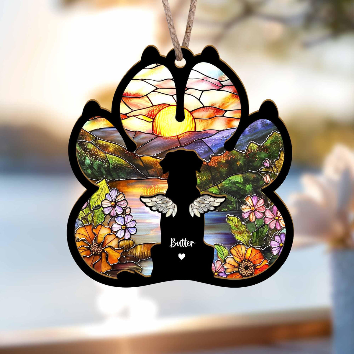 May include: A wooden paw print ornament with a stained glass design of a sunset over mountains and a dog with angel wings. The dog is silhouetted and the ornament is personalized with the name "Butter".